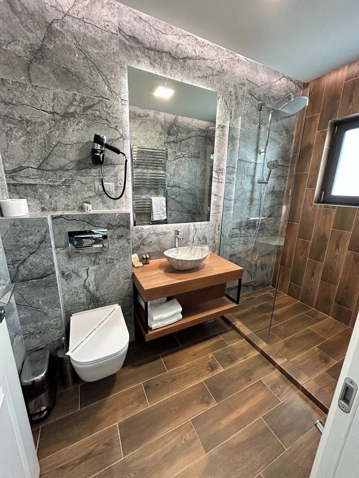 A bathroom with a marble wall, a wooden vanity, a white toilet, and a glass-enclosed shower area.