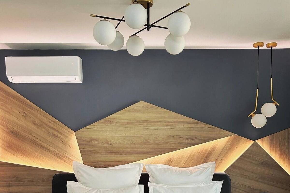 A modern bedroom with a geometric wooden headboard, a central bed with white bedding and black pillows, and a ceiling light fixture with multiple spherical bulbs.