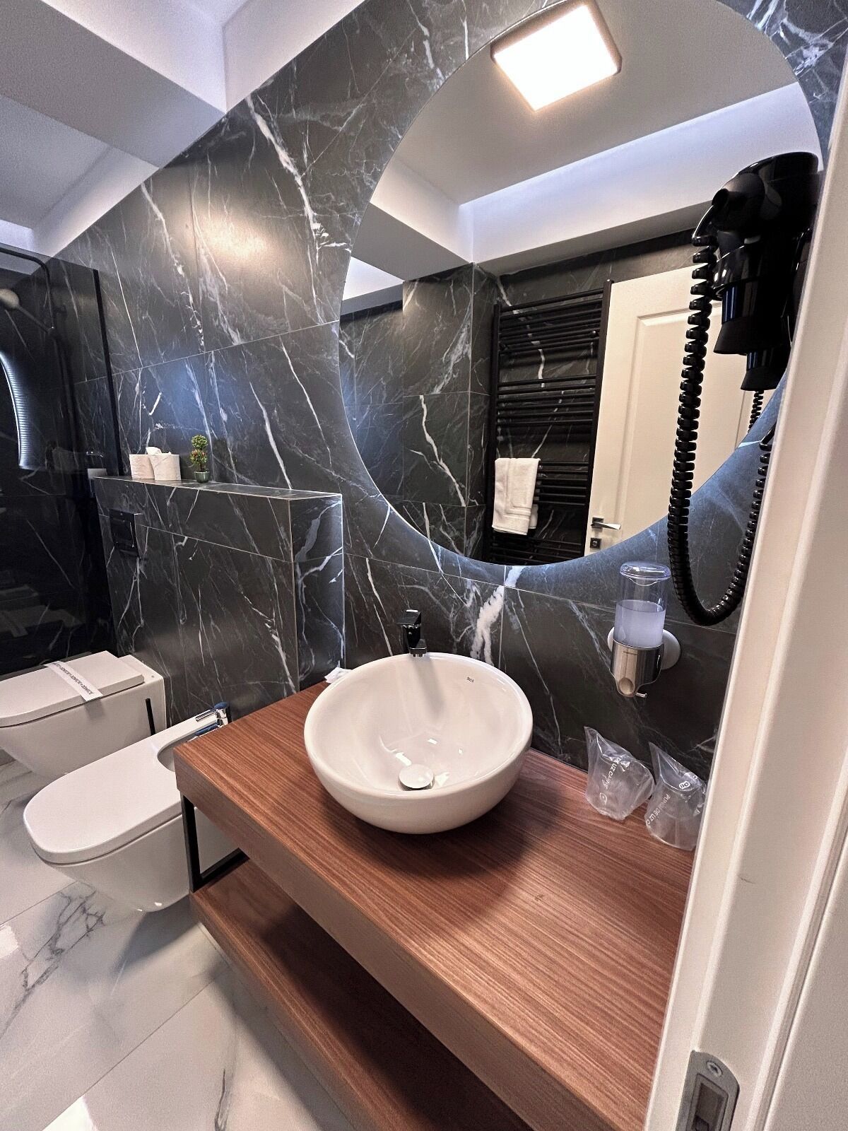 A modern bathroom with a marble backsplash, a wooden vanity, a white sink, a toilet, and a hairdryer mounted on the wall.