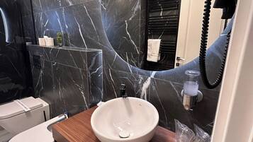 A modern bathroom with a marble backsplash, a wooden vanity, a white sink, a toilet, and a hairdryer mounted on the wall.