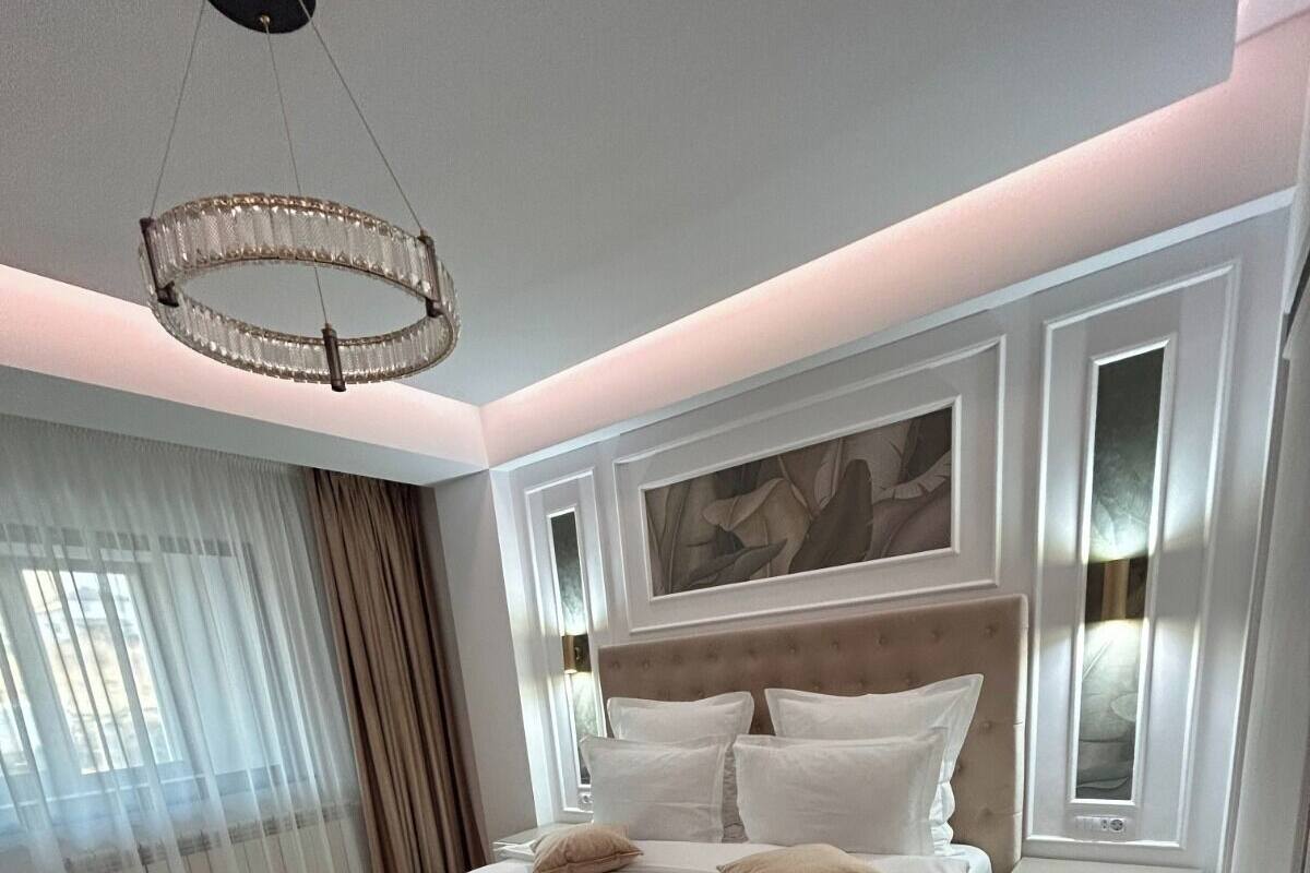 A modern bedroom with a large bed, a headboard with built-in lighting, a bedside table, and a large window with sheer curtains.