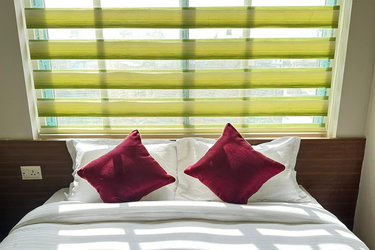A bed with white bedding and two red pillows, positioned in front of a window with horizontal blinds.
