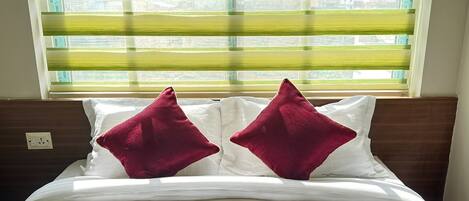 Una cama con ropa de cama blanca y dos almohadas rojas, frente a una ventana con persianas horizontales.
