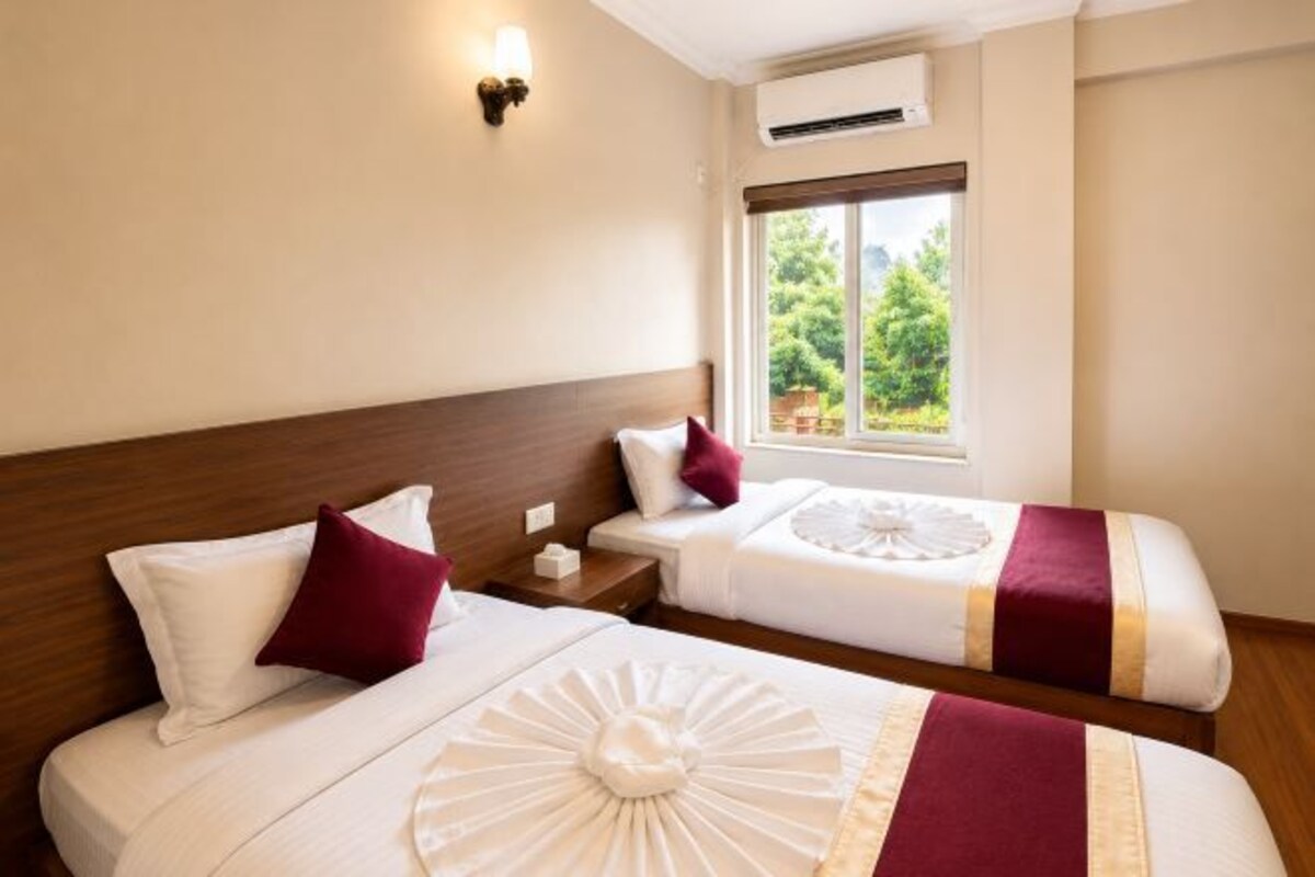 A hotel room with two beds, a wooden headboard, a window with a view of greenery, and an air conditioning unit.