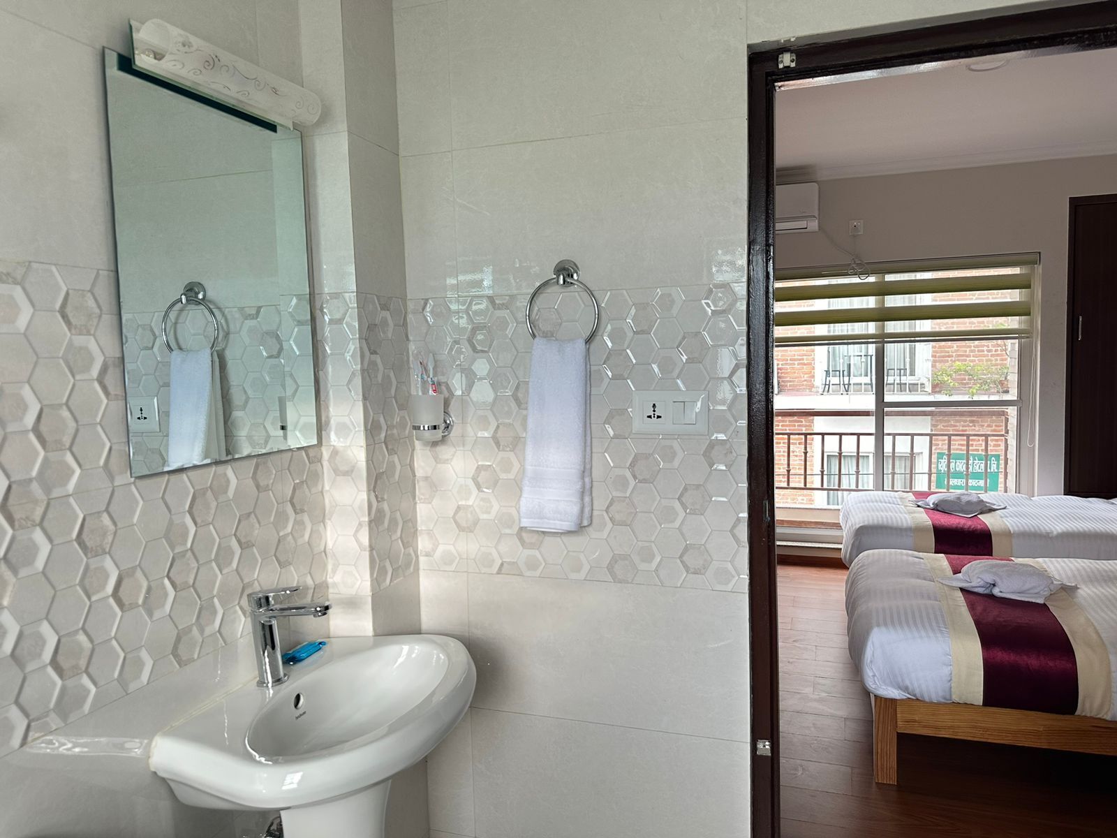 A bathroom with a sink, mirror, and towel rack. A bed is visible in the adjacent room.