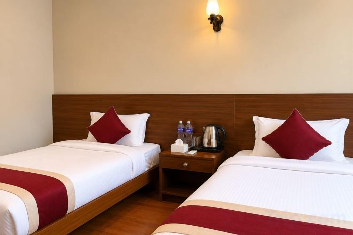 A hotel room with two beds, each with a red pillow and a wooden headboard. There is a nightstand with a kettle and bottles.