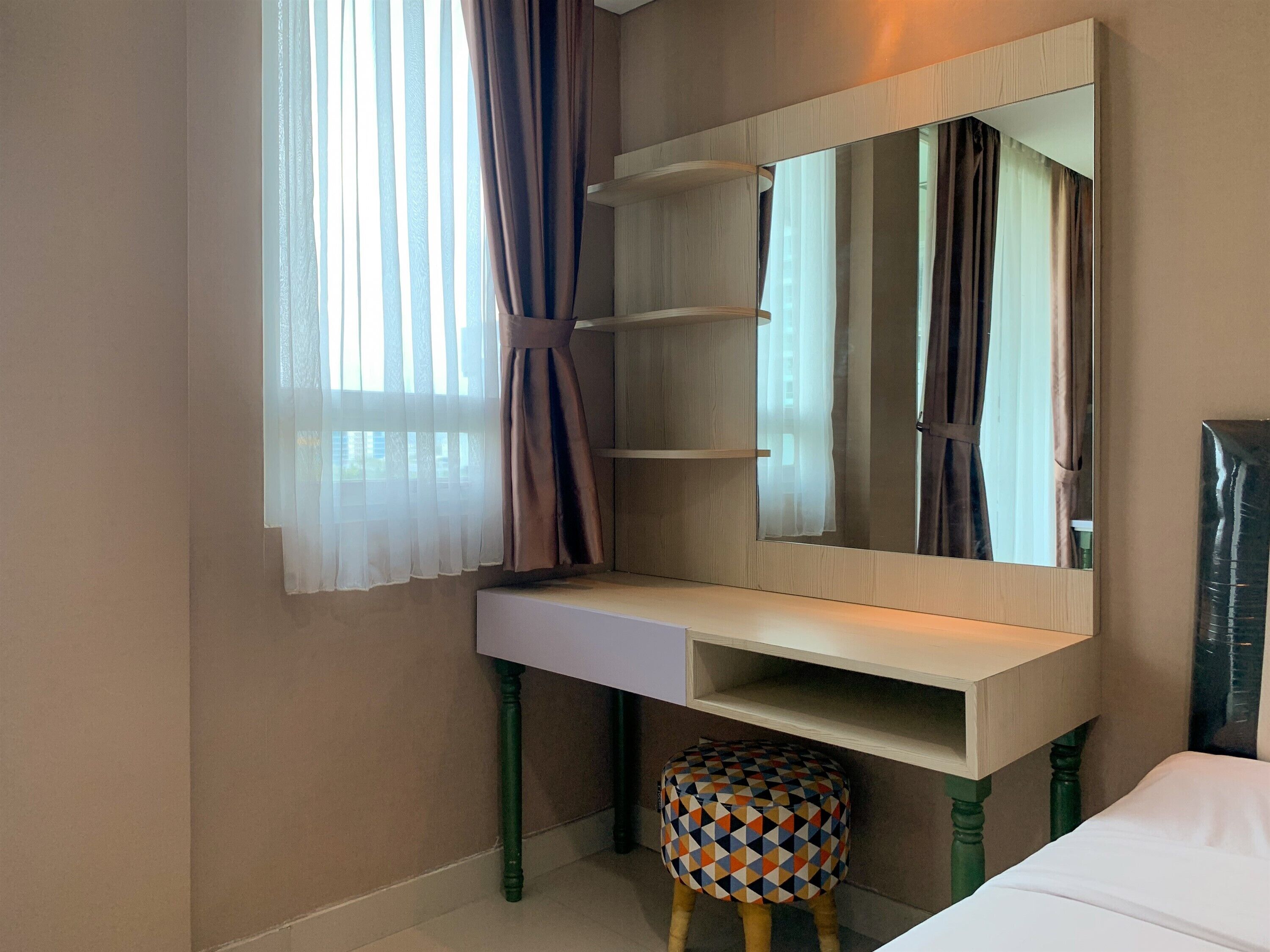 A bedroom with a bed, a bedside table, a mirror, a window with curtains, and a small stool.