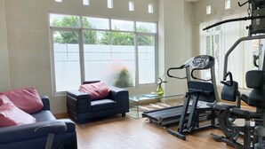 Un gimnasio en casa con cinta de correr, banco para pesas y equipo de ejercicio. Se ve una ventana grande con vistas a árboles y una cerca blanca. También se observa una silla con un cojín rosa.