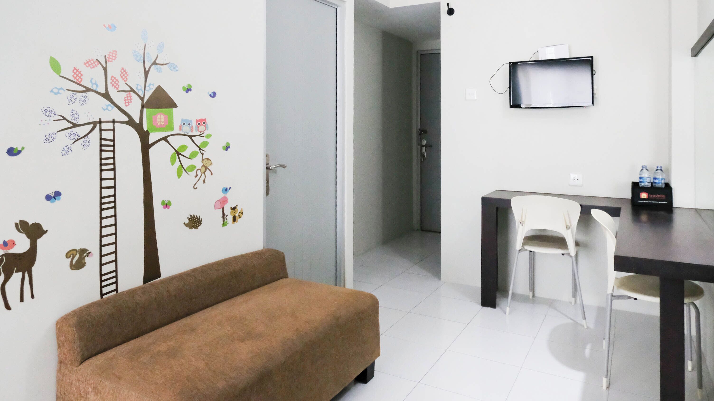 A room with a brown sofa, a dining table with chairs, a TV mounted on the wall, and a wall decal featuring a tree with animals.