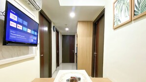 Quarto de hotel com televisão na parede exibindo diversos serviços de streaming, uma mesa de jantar de madeira com um jogo americano floral e um sofá castanho.