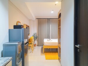 Una habitación de hotel compacta con cama, un escritorio pequeño con silla, refrigerador y una planta en maceta.