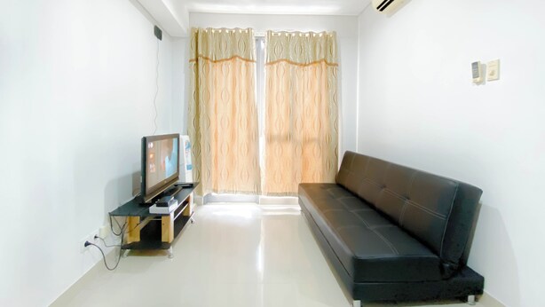 A living room with a black sofa, a television on a wooden stand, and a white air conditioner on the wall. - Homey and Modern 2BR Apartment at Royal Olive Residence (Jakarta)