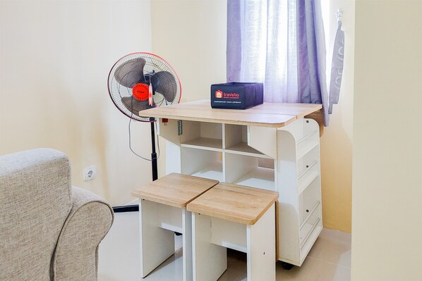 Une pièce de taille modeste, avec un bureau en bois, un petit ventilateur et une valise de voyage.