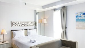 Une chambre d’hôtel avec un lit, une table de chevet avec une lampe, une chaise, un tableau représentant une plage et une fenêtre avec des rideaux.