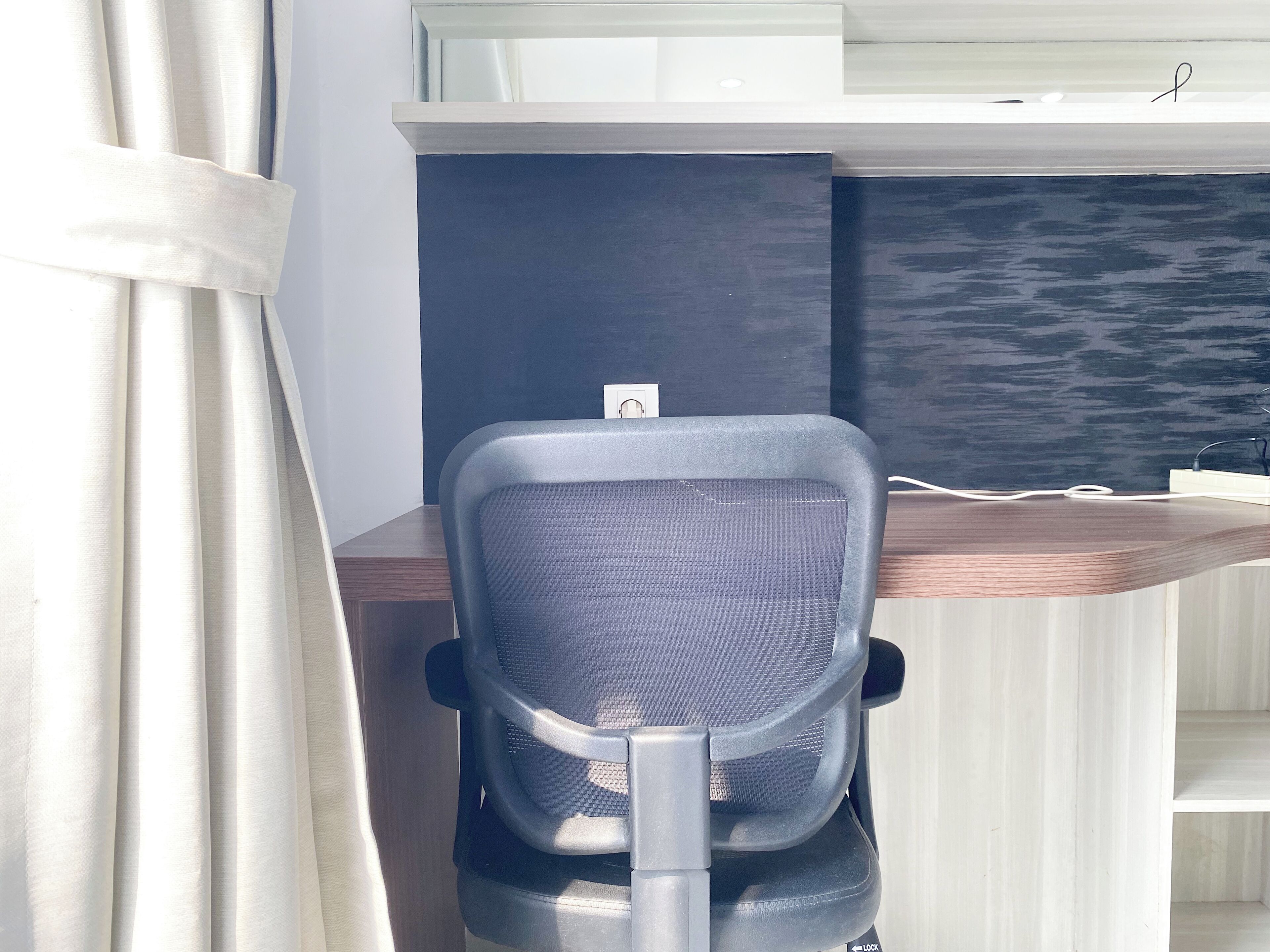 A modern office chair with a mesh backrest and armrests, positioned in front of a desk with a dark back panel and a white cable running along the edge.