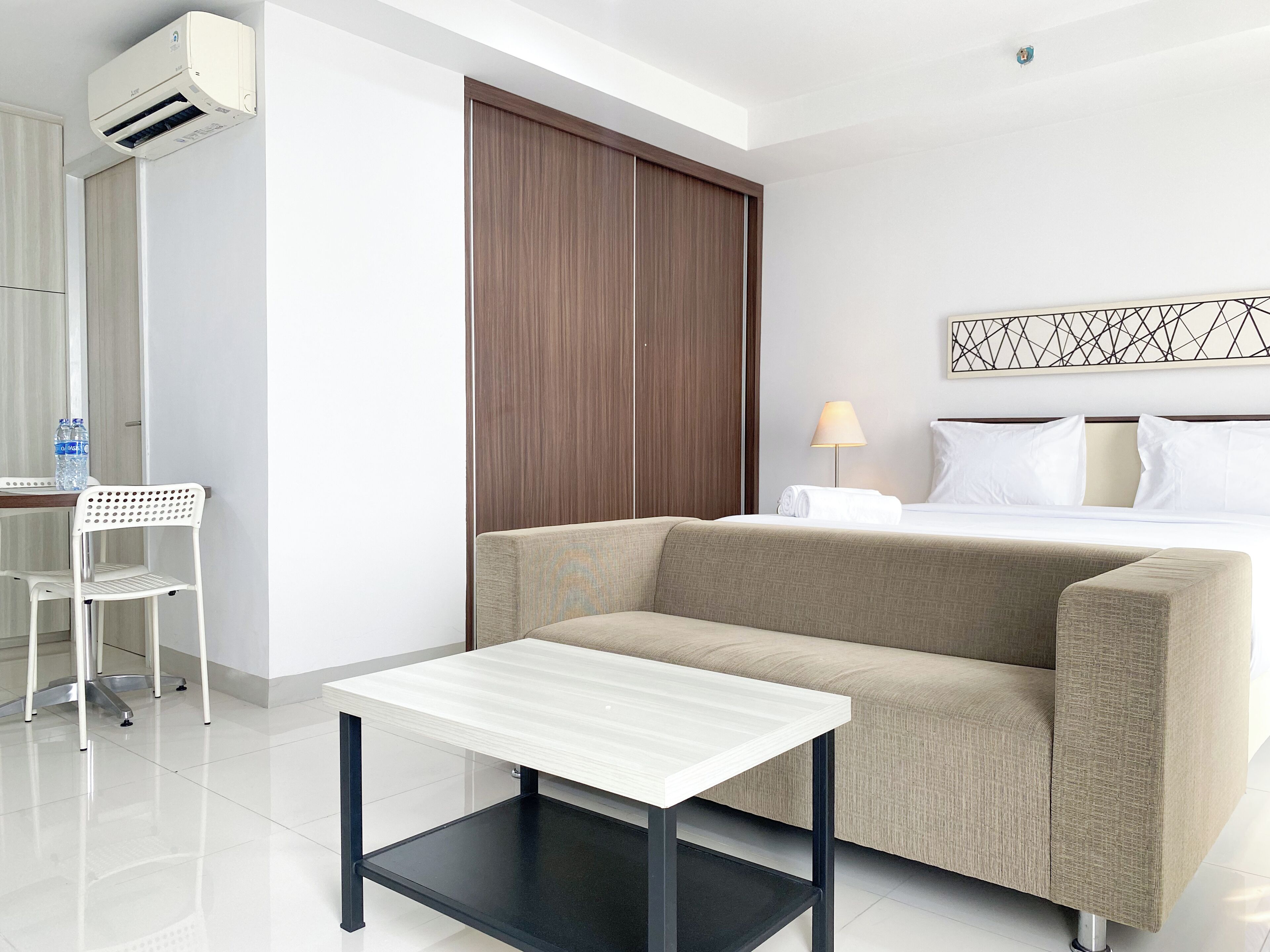 A hotel room with a sofa, a small table, a chair, a bed, a nightstand, a lamp, and an air conditioning unit.