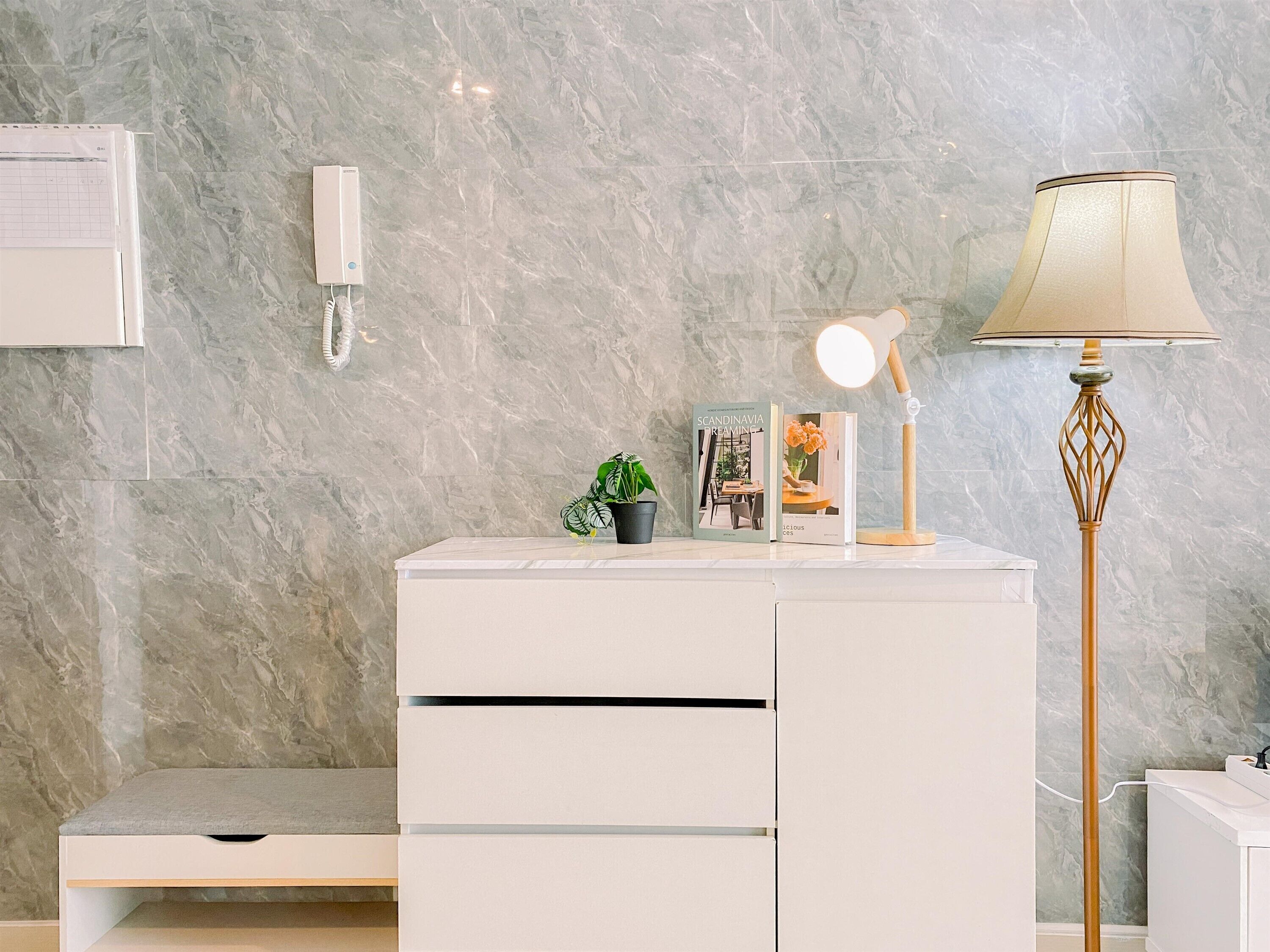 Een moderne badkamer met een marmeren wand, een witte wastafel met lades, een kleine potplant, een tijdschrift, een lamp en een telefoon aan de muur.