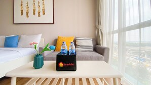 Een moderne hotelkamer met een witte bank, een klein tafeltje met waterflesjes, een vaas met een bloem en een lijst met een kunstwerk aan de muur.