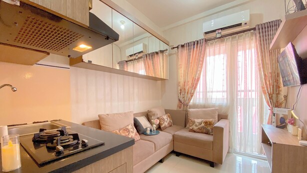 A modern kitchen with a built-in stove, a sink, and a countertop. There is a beige sofa with decorative pillows and a window with sheer curtains. - Good and Warm 2BR at Green Pramuka City Apartment (Jakarta)