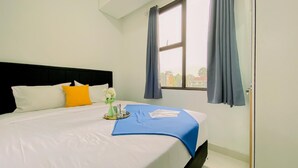A neatly made bed with white and yellow pillows, a blue bedspread, and a small table with a vase of flowers and a tray with toiletries. - Brand New and Homey 2BR at Transpark Cibubur Apartment (Depok)