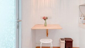 Une salle de bain avec un lavabo blanc, une étagère en bois, un tabouret blanc, une poubelle et un luminaire en osier.