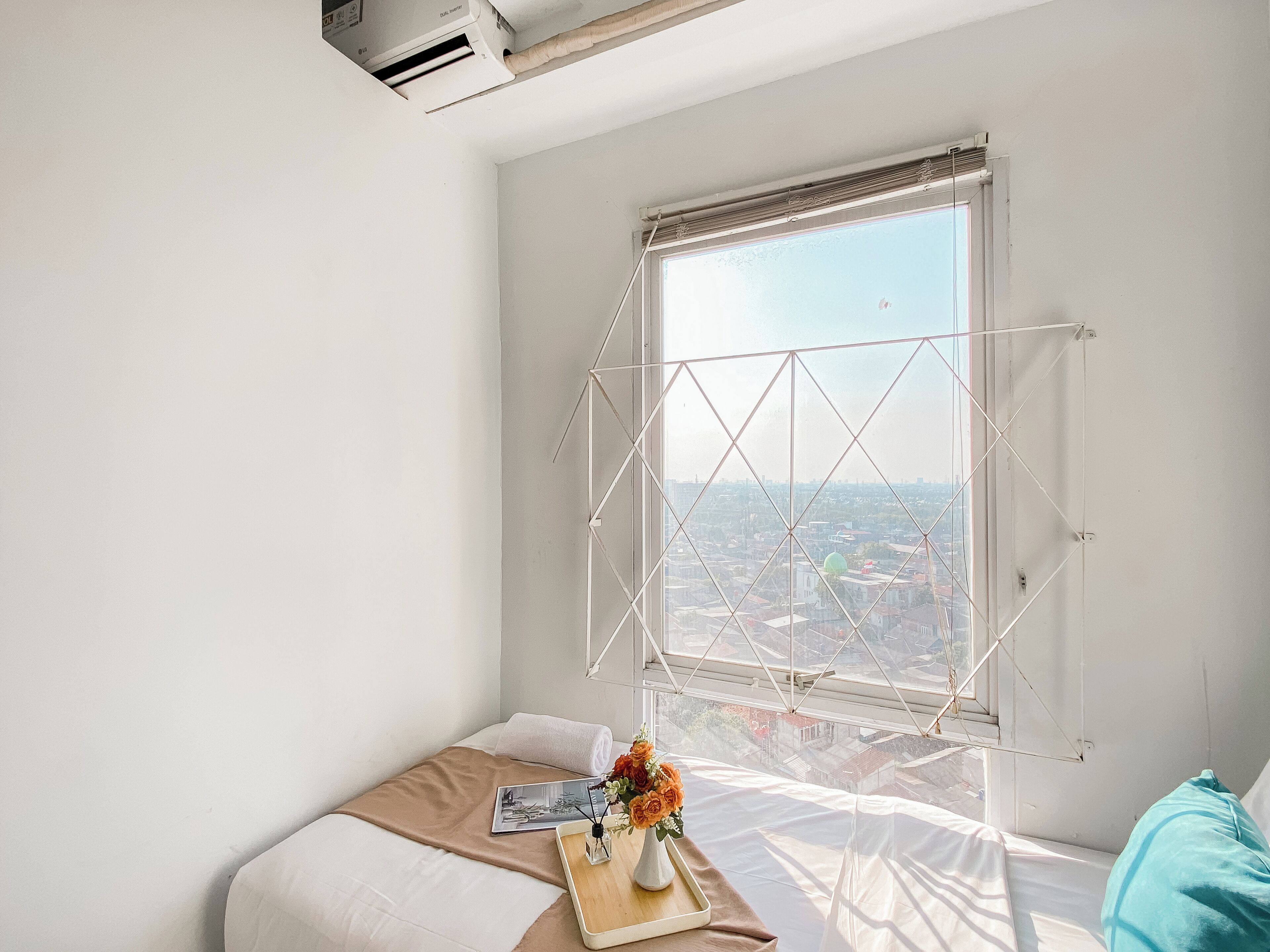 A bedroom with a large window, a bed, a bedside table with a vase of flowers, and a view of a cityscape.