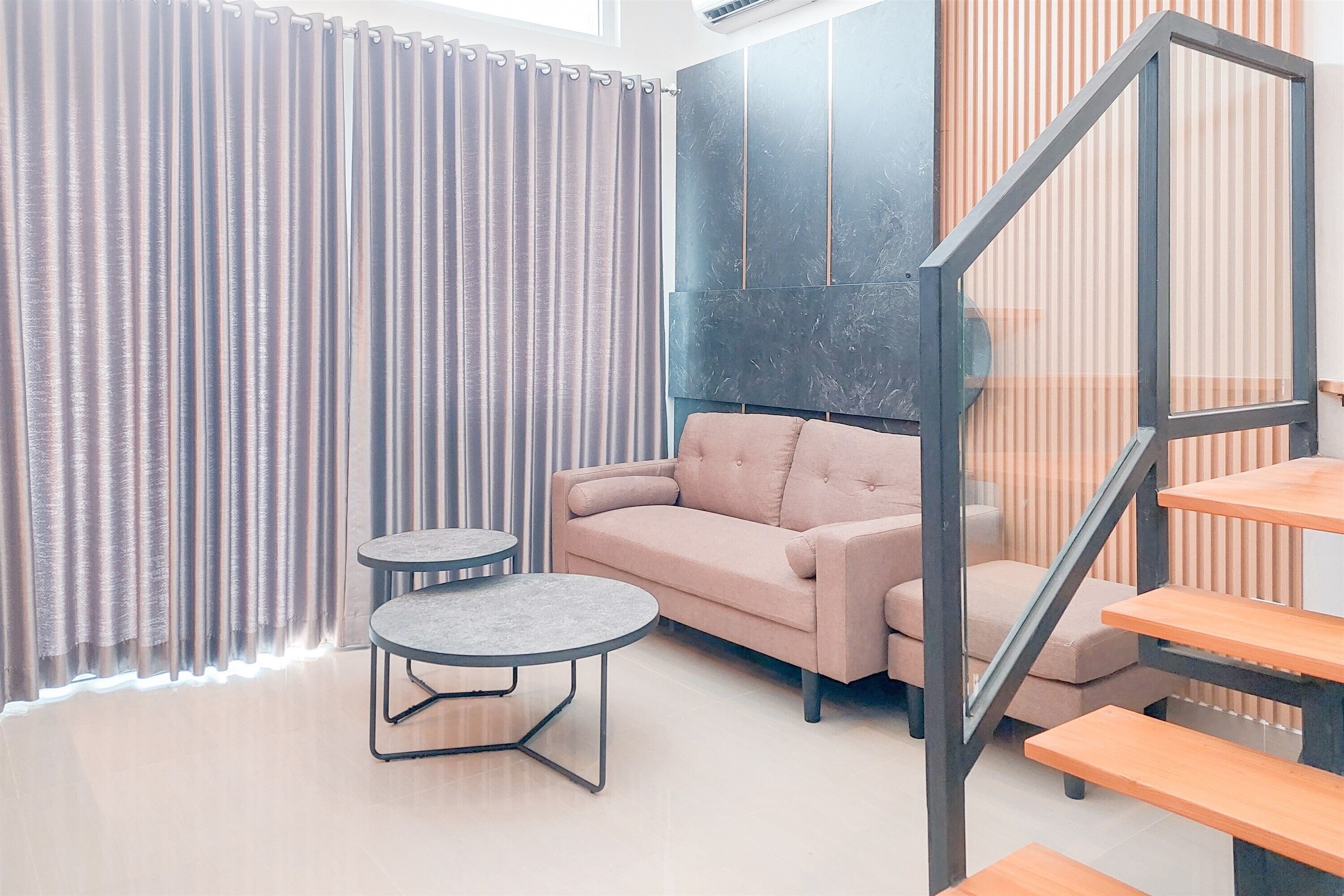 Una sala de estar moderna con un sofá, dos mesitas, una escalera y un ventanal con cortinas.