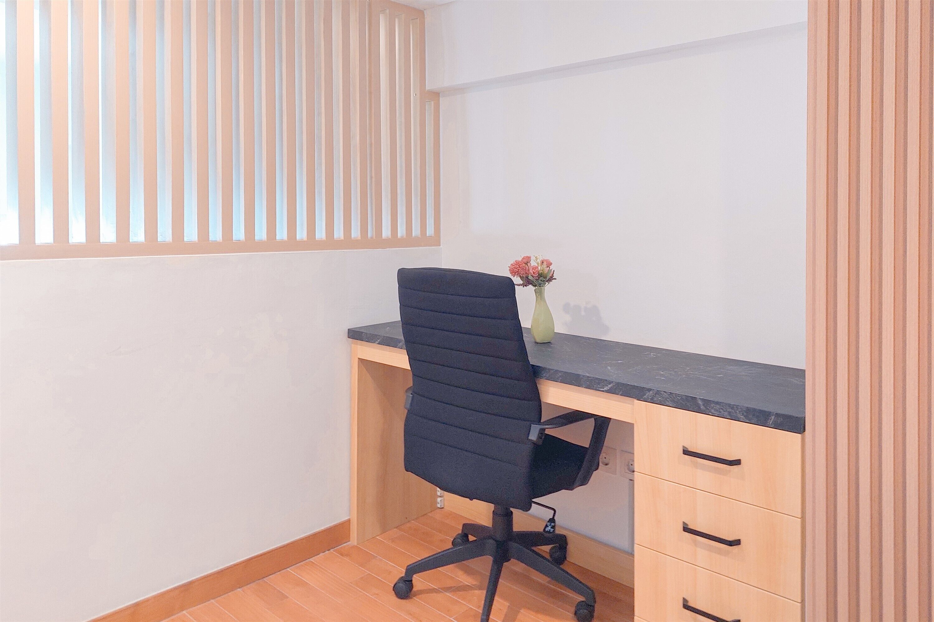 Une pièce comprenant un bureau, une chaise, un vase avec des fleurs et une fenêtre avec des stores.