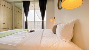 A neatly made bed with white linens, a bedside table with a lamp, and a window with curtains. - Enjoy Living Studio at West Vista Apartment (Jakarta)