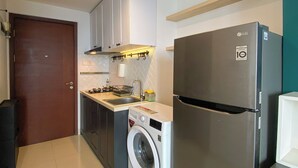 A compact kitchen with a washing machine, a refrigerator, and a wooden door. - Enjoy Living Studio at West Vista Apartment (Jakarta)