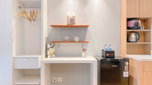 A compact kitchen with a refrigerator, a kettle, and a shelf with a travel bag.