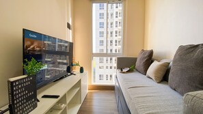 A living room with a flat-screen TV on a stand, a sofa, and a window with a view of a building.