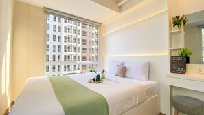 A hotel room with a large bed, a bedside table with a plant, a wall-mounted shelf with a sign listing cities, and a window with a view of a building.