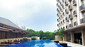 A swimming pool with a checkered blue tile pattern, surrounded by buildings with balconies and a covered area with seating. - Modern Look and Restful Studio at Azalea Suites Apartment (Cikarang)