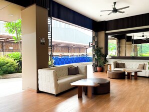 Un salon moderne comprenant un canapé, un pouf et une table basse, une grande fenêtre donnant sur une piscine et un espace de détente extérieur, ainsi qu’un ventilateur de plafond.