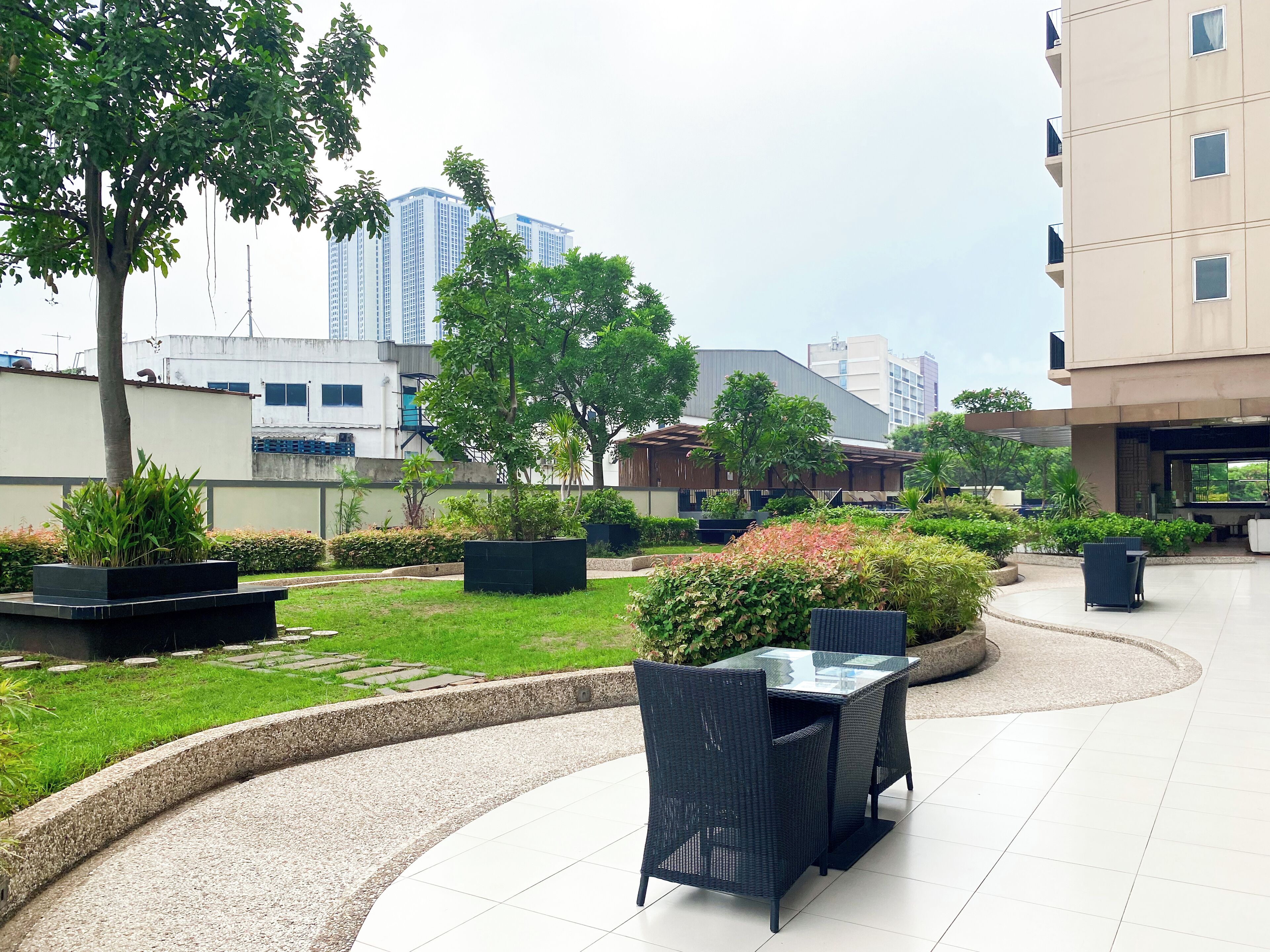 An outdoor patio area with a curved seating arrangement, surrounded by greenery and a view of high-rise buildings in the background.