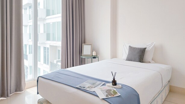 A neatly made bed with a blue blanket, a small table with a candle, and a window with curtains. - Good Choice and Restful 1BR at Tree Park City BSD Apartment (South Tangerang)