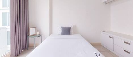 A minimalist hotel room with a bed, a small bedside table with a lamp, a window with curtains, and a white wardrobe.