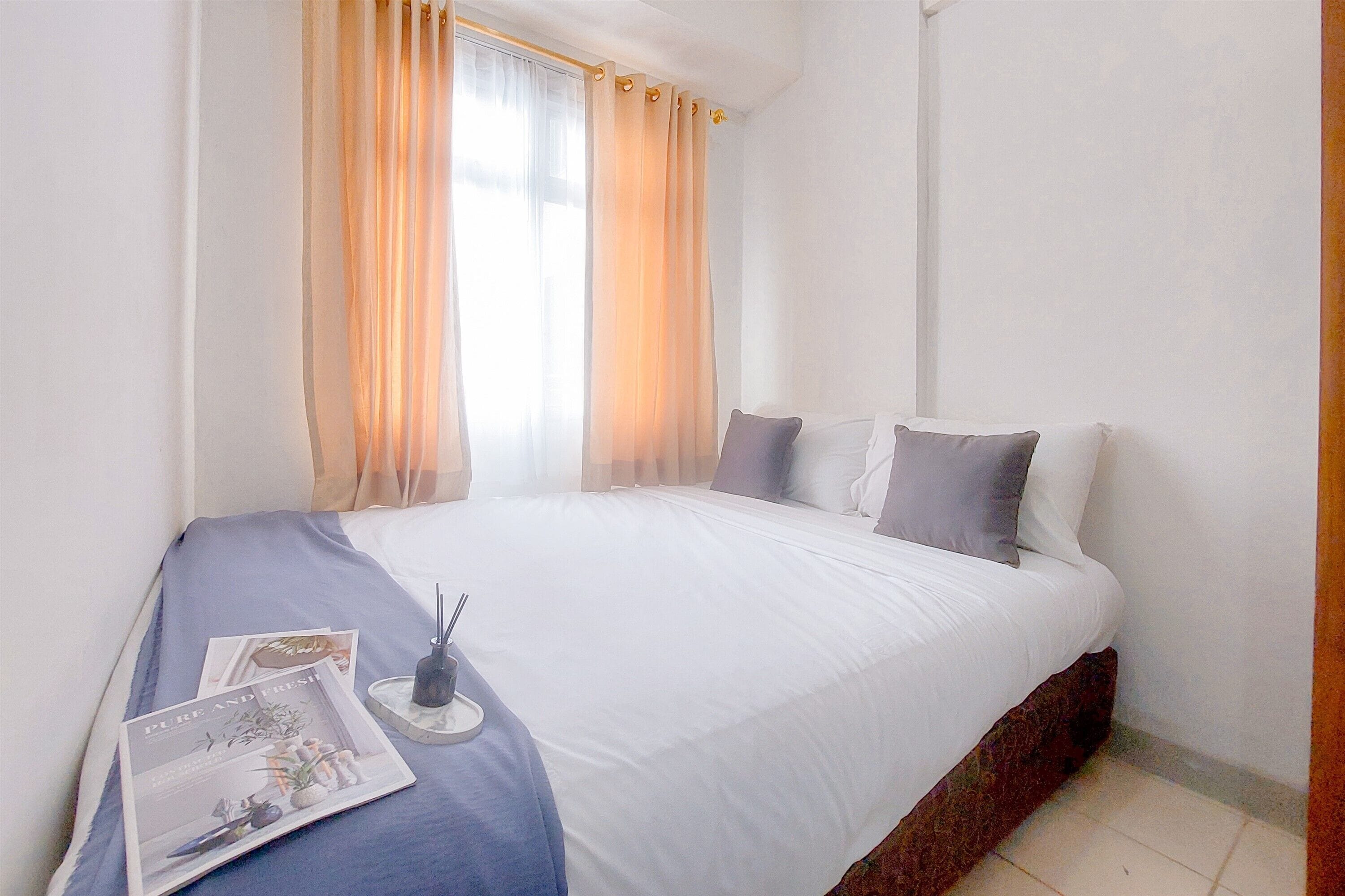 A neatly made bed with white linens, a bedside table with a lamp, and a window with sheer curtains.