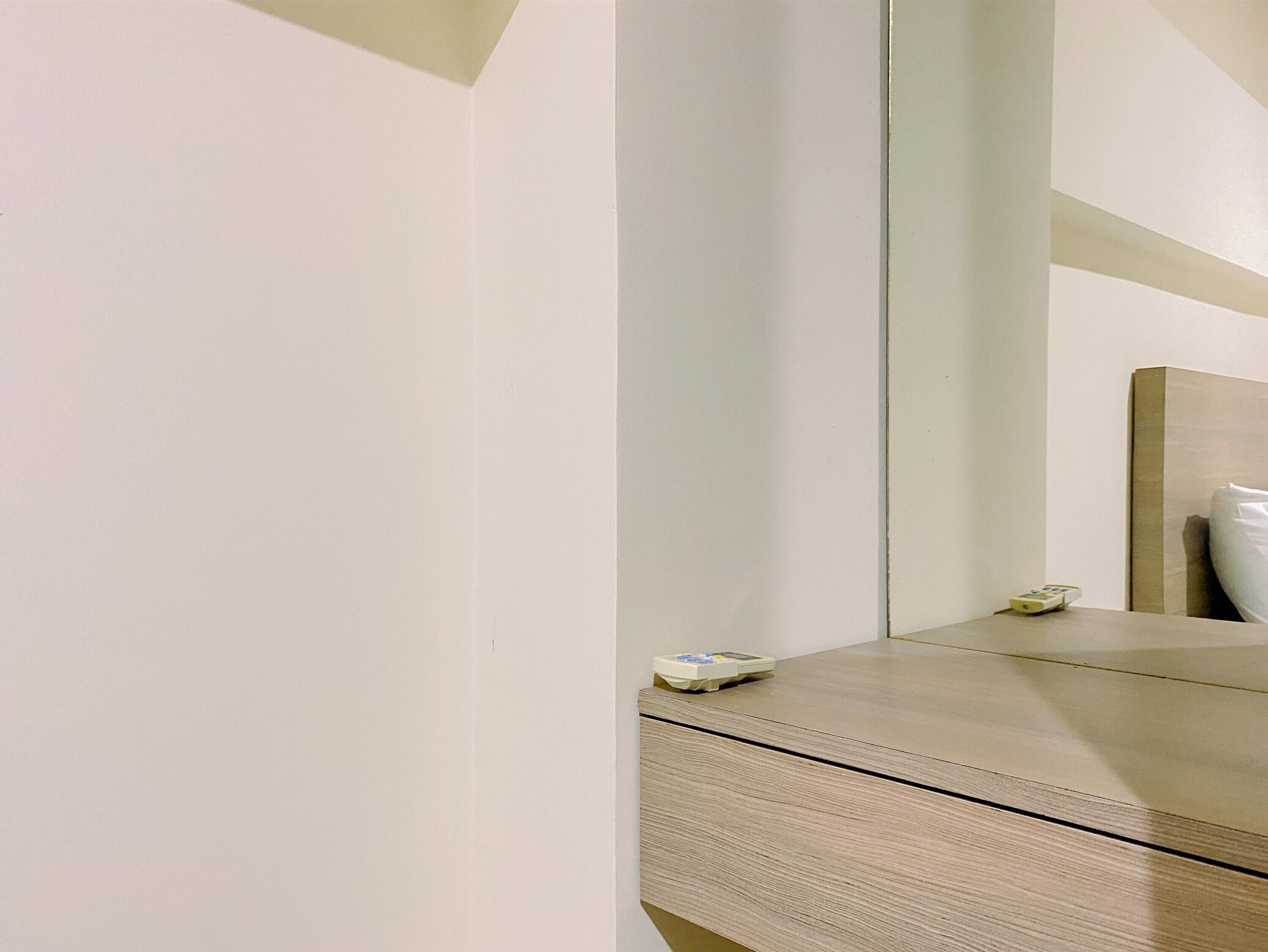 Ein schlicht eingerichtetes Hotelzimmer mit Nachttisch aus Holz, einem kleinen Beistelltisch und einem Spiegel, der das Licht an den hellen Wänden und dem Bett mit weißer Bettwäsche reflektiert.