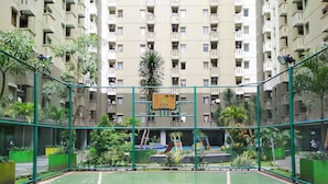 Un campo da basket all'aperto circondato da una recinzione verde, con un'area giochi sullo sfondo e un edificio residenziale con numerosi balconi.