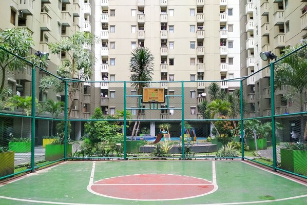 Un terrain de basket en plein air, entouré d’une clôture verte, avec une aire de jeux en arrière-plan et un immeuble d’habitation doté de nombreux balcons.