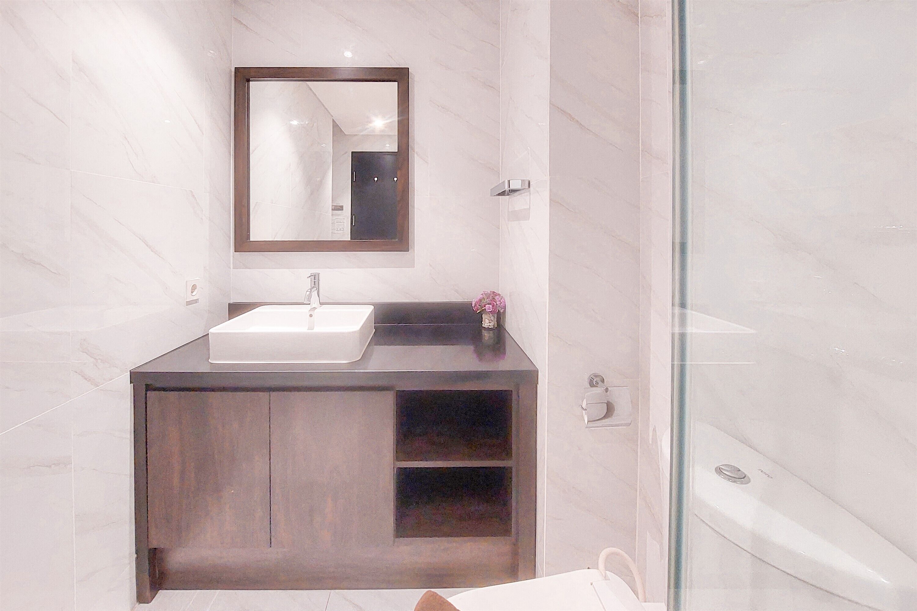 A modern bathroom with a marble wall, a wooden vanity, a white sink, a mirror, and a glass shower enclosure.