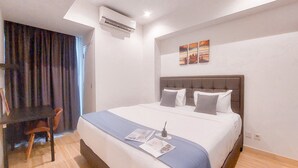 A hotel room with a large bed, a desk, and a chair. There is an air conditioning unit on the wall and a window with curtains. - Good Deal and Restful 1BR at Branz BSD City Apartment (Pagedangan)
