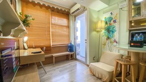 Habitación luminosa con escritorio, silla y banco. Amplia ventana con persianas, dispensador de agua y un cuadro en la pared.