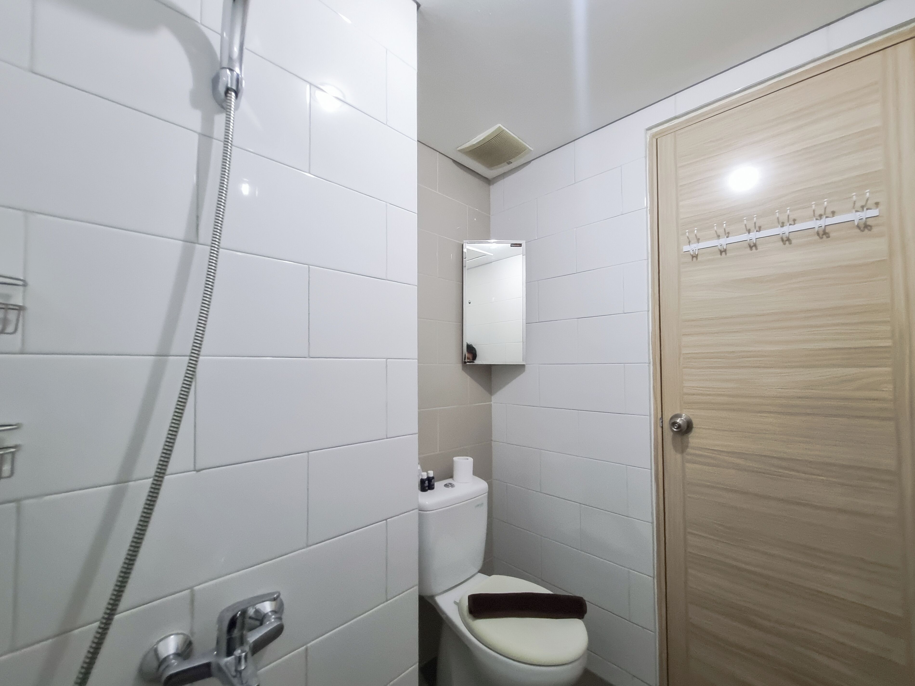 A small, clean bathroom with white tiles, a shower, a toilet, and a wooden door.
