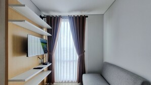 A modern living room with a flat-screen TV mounted on a wooden shelf, a grey sofa, and a large window with curtains. - Cozy Living Studio at Taman Melati Sinduadi Apartment (Pogung Lor)