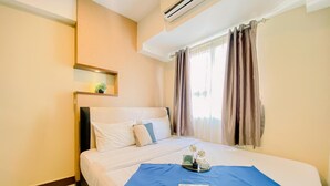 A hotel room with a bed, a bedside table with a lamp, a window with curtains, and an air conditioning unit. - Homey and Comfort Stay 2BR Cinere Resort Apartment (Depok)