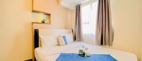 A hotel room with a bed, a bedside table with a lamp, a window with curtains, and an air conditioning unit.