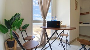 Une petite pièce avec une table, deux chaises, une plante en pot, une fenêtre avec des rideaux et une vue sur un paysage lointain.